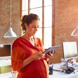 Article thumbnail: Pregnant businesswoman using tablet PC. Female professional is standing at desk. She is in brightly lit office.