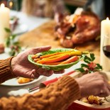 Article thumbnail: Christmas dinner being served on table with vegetables and serving dish of roast potatoes, roast turkey resting in background, lit candles and red wine